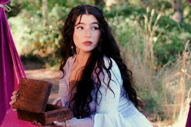 Musician Effie Isobel posing outdoors in a lavender dress, looking over her shoulder while opening a small wooden treasure box.
